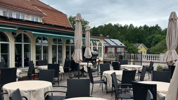 Eine Terrasse im Freien mit runden Tischen und Stühlen, beschattet durch Sonnenschirme, und ein Gebäude mit Ziegeldach im Hintergrund.