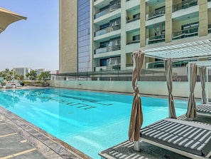 A swimming pool with a clear blue water surface, surrounded by lounge chairs and umbrellas, with a modern building in the background. - Stunning 1BR, 8 Min to Burj Khalifa (Dubai)