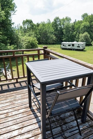 A wooden deck with a table and chair, a railing, and a green landscape with trees and a trailer in the background.