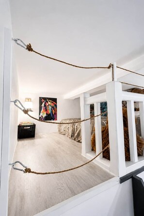 Una habitación con techo blanco, suelo de madera, un cuadro abstracto colorido, una cesta negra y una cama blanca con una manta marrón.