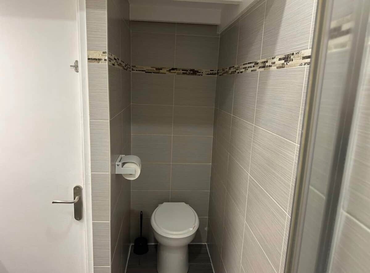 Een kleine, smalle badkamer met een toilet, een toiletrolhouder en een bedieningsknop voor het doorspoelen.