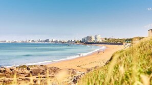 Un paysage urbain côtier avec une plage de sable, une eau bleue et limpide, et des bâtiments visibles au loin.