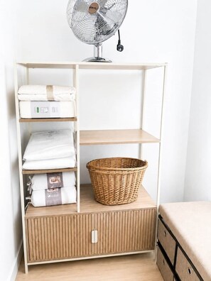 Un armario blanco con una cesta de mimbre, toallas dobladas y un ventilador de pie.