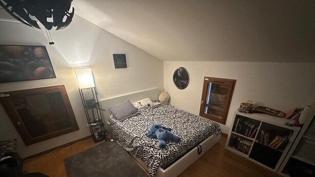 Et soveværelse med seng, skrivebord, stol, hylde med bøger og en lampe på væggen.