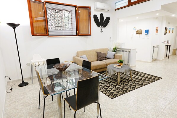 Un salon avec une table à manger en verre, des chaises noires, un canapé beige, un tapis à motifs noir et blanc et un climatiseur fixé au mur.