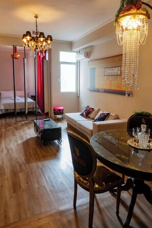 A hotel room with a bed, a sofa, a dining table with chairs, a chandelier, and a window. - Monastiraki Art House (Athina)