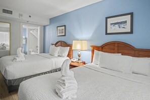 Quarto de hotel com duas camas, mesa de cabeceira com luminária, quadro na parede e porta de acesso a um banheiro.
