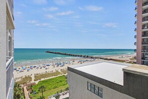 A beach view with a pier extending into the ocean, a sandy beach with umbrellas, and a building with balconies overlooking the scene. - Oceanview 1BR Suite + Lazy River & Hot Tub (Myrtle Beach)