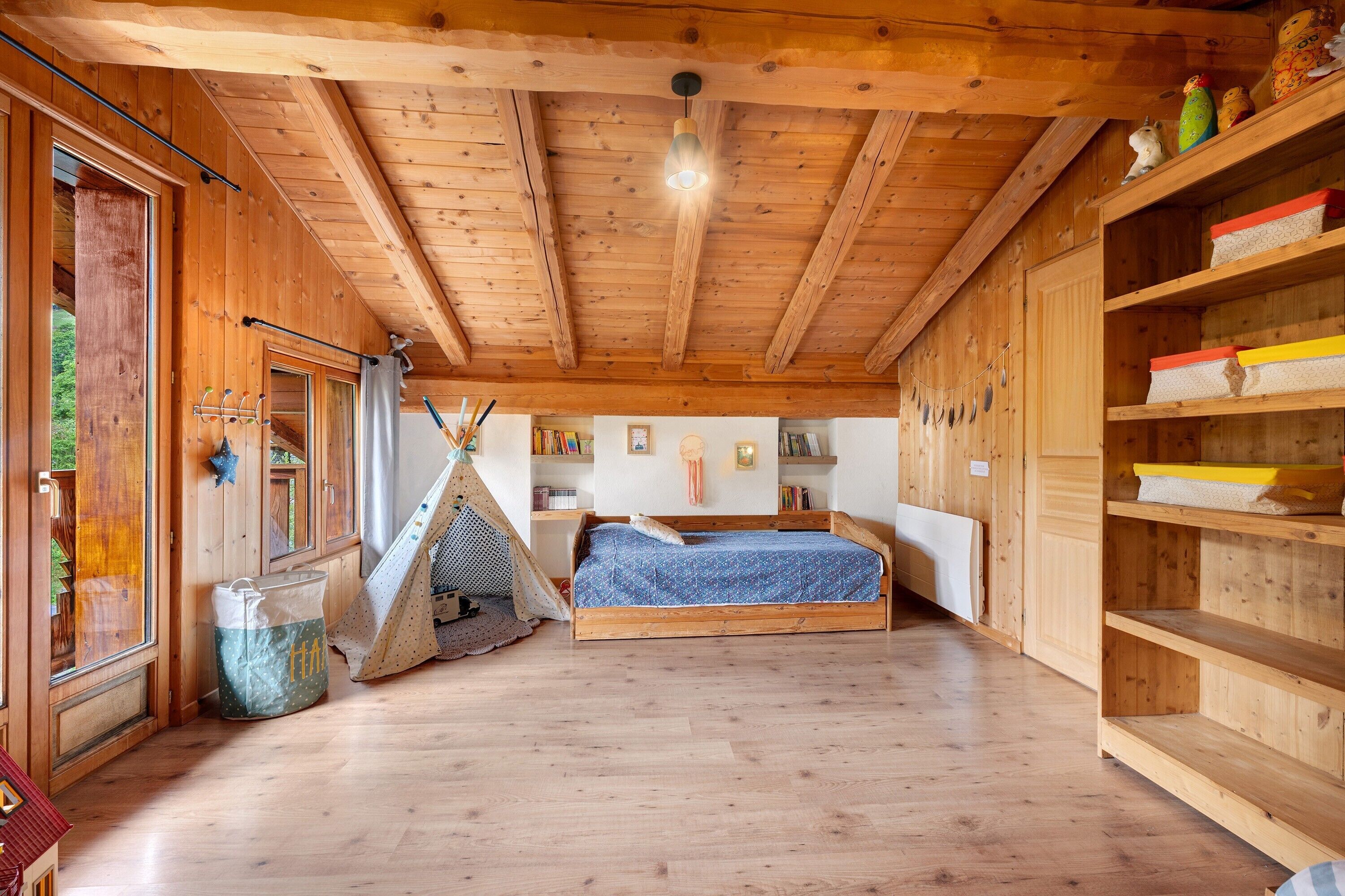 Ein Holzinnenraum mit einem Bett, einem Regal mit Aufbewahrungsboxen und einem Fenster mit Vorhängen.