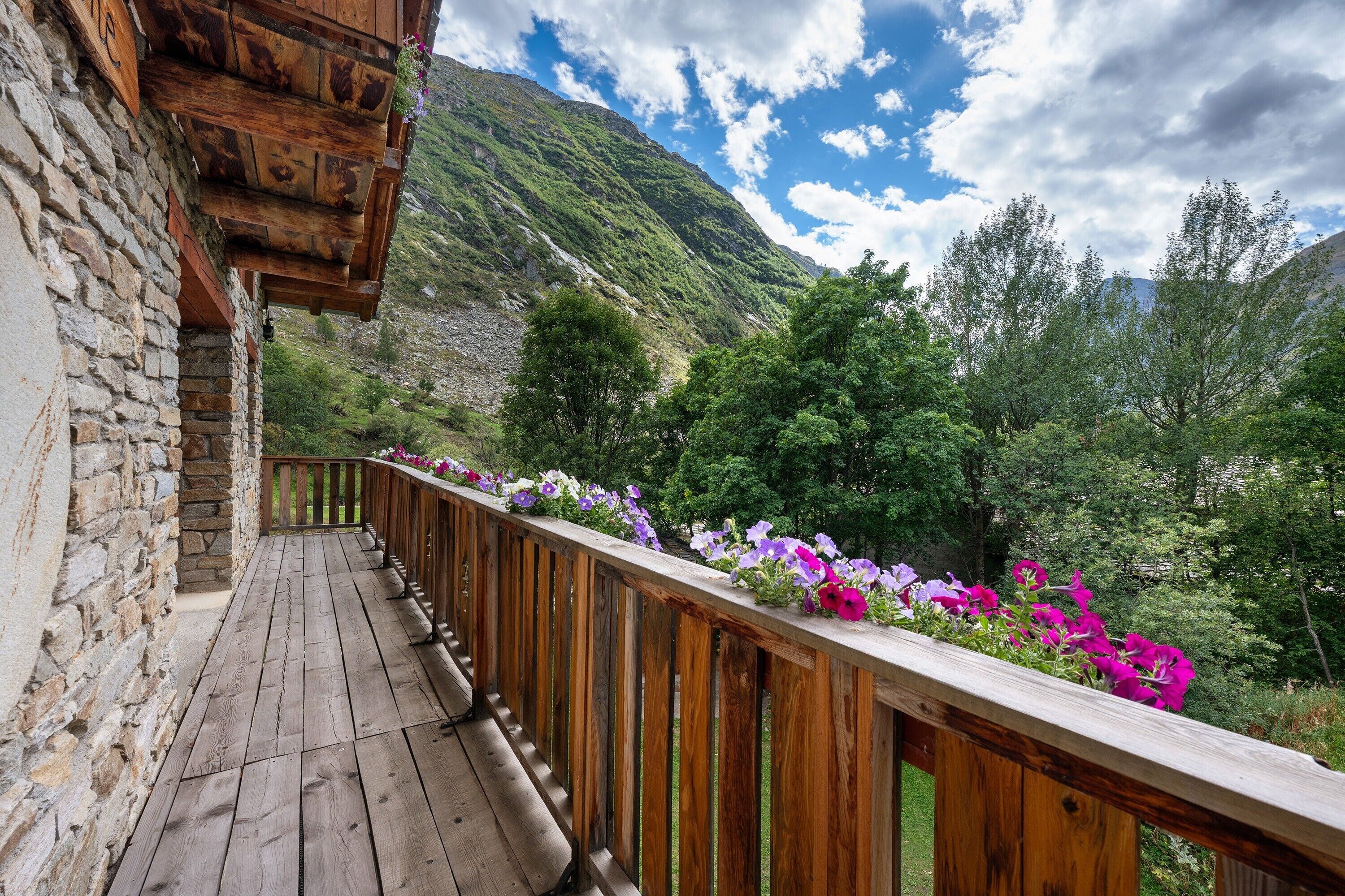 A wooden deck with a stone wall, overlooking a mountainous landscape with flowering plants.