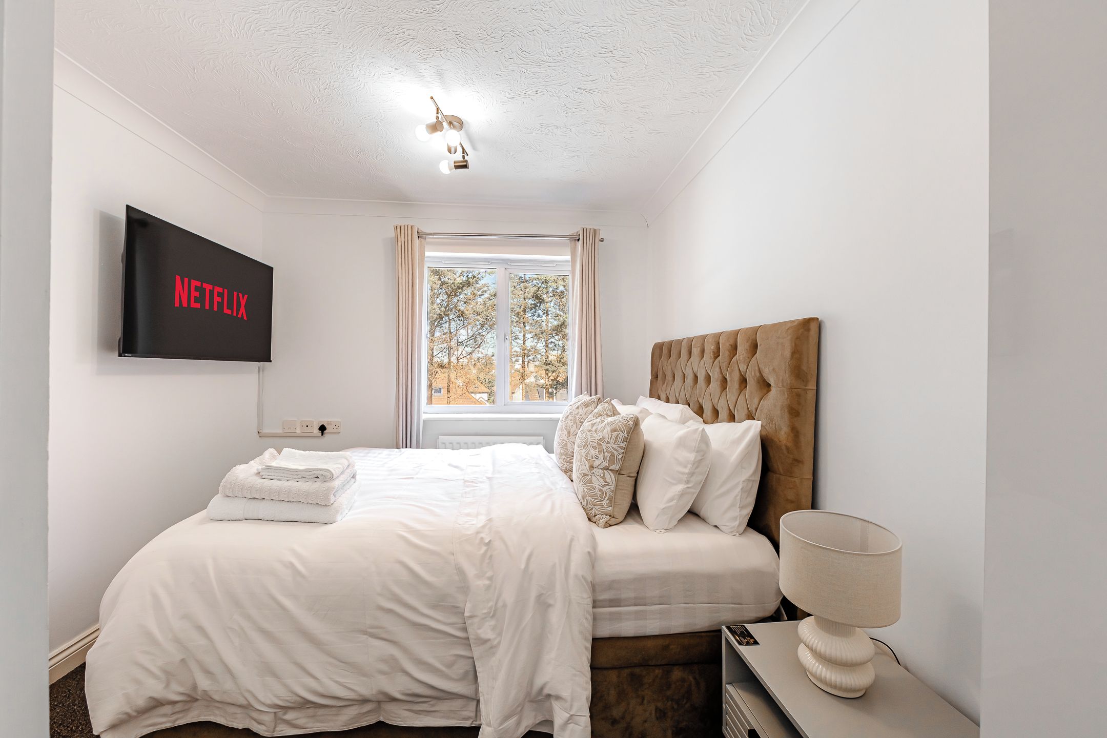 A bedroom with a bed, a TV mounted on the wall, a window with curtains, and a bedside lamp.