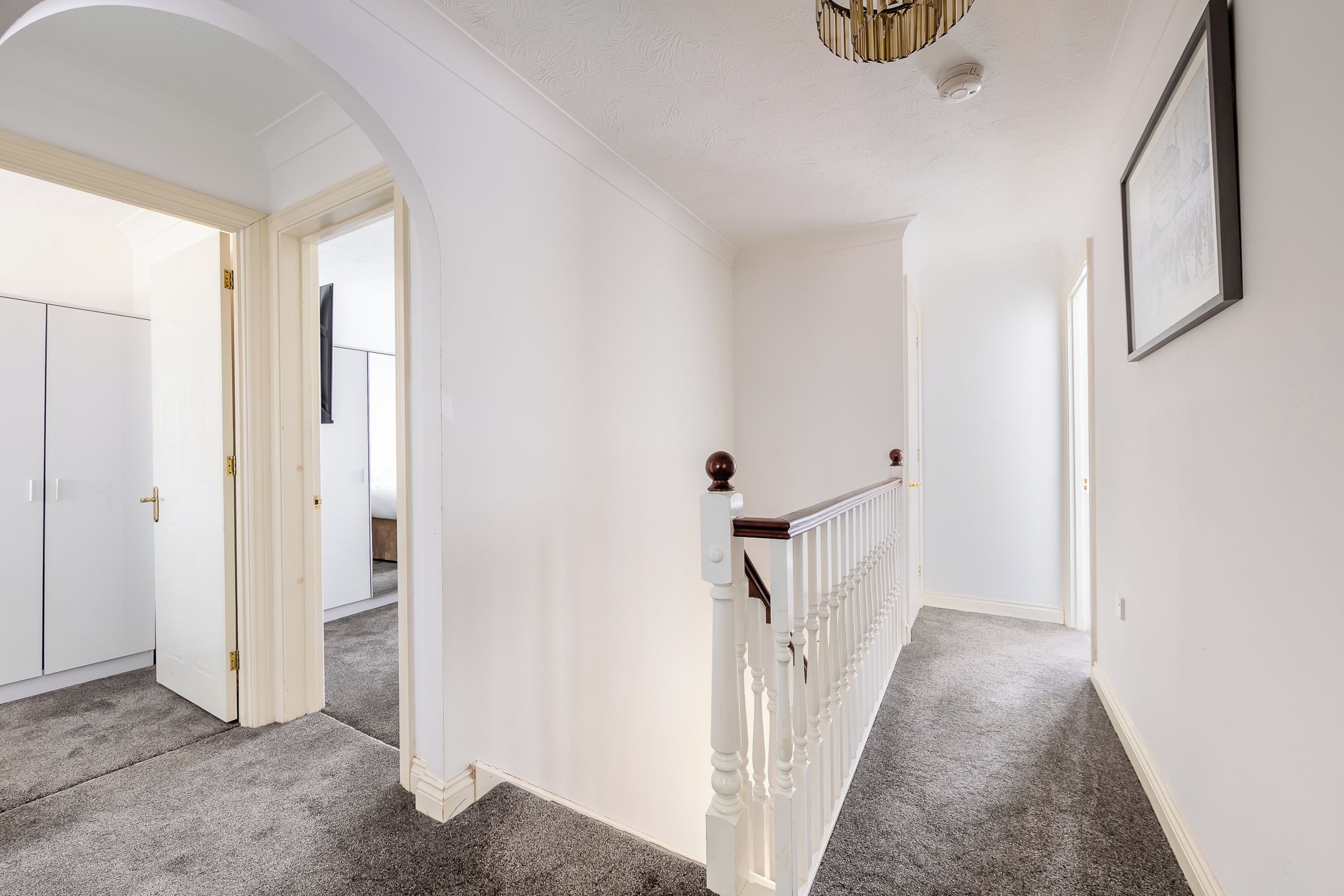 A hallway with a carpeted floor, white walls, a wooden railing, and a door leading to another room.