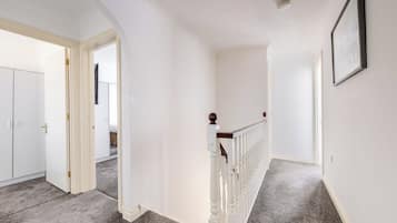 A hallway with a carpeted floor, white walls, a wooden railing, and a door leading to another room.