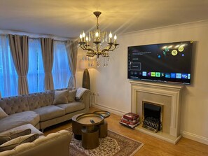 A living room with a large flat-screen TV mounted above a fireplace, a sofa, a coffee table, and a chandelier. - Luxury 4 Bedroom Home, Sleeps 10, Garden, Free Parking, Hornchurch, Near London (Hornchurch)