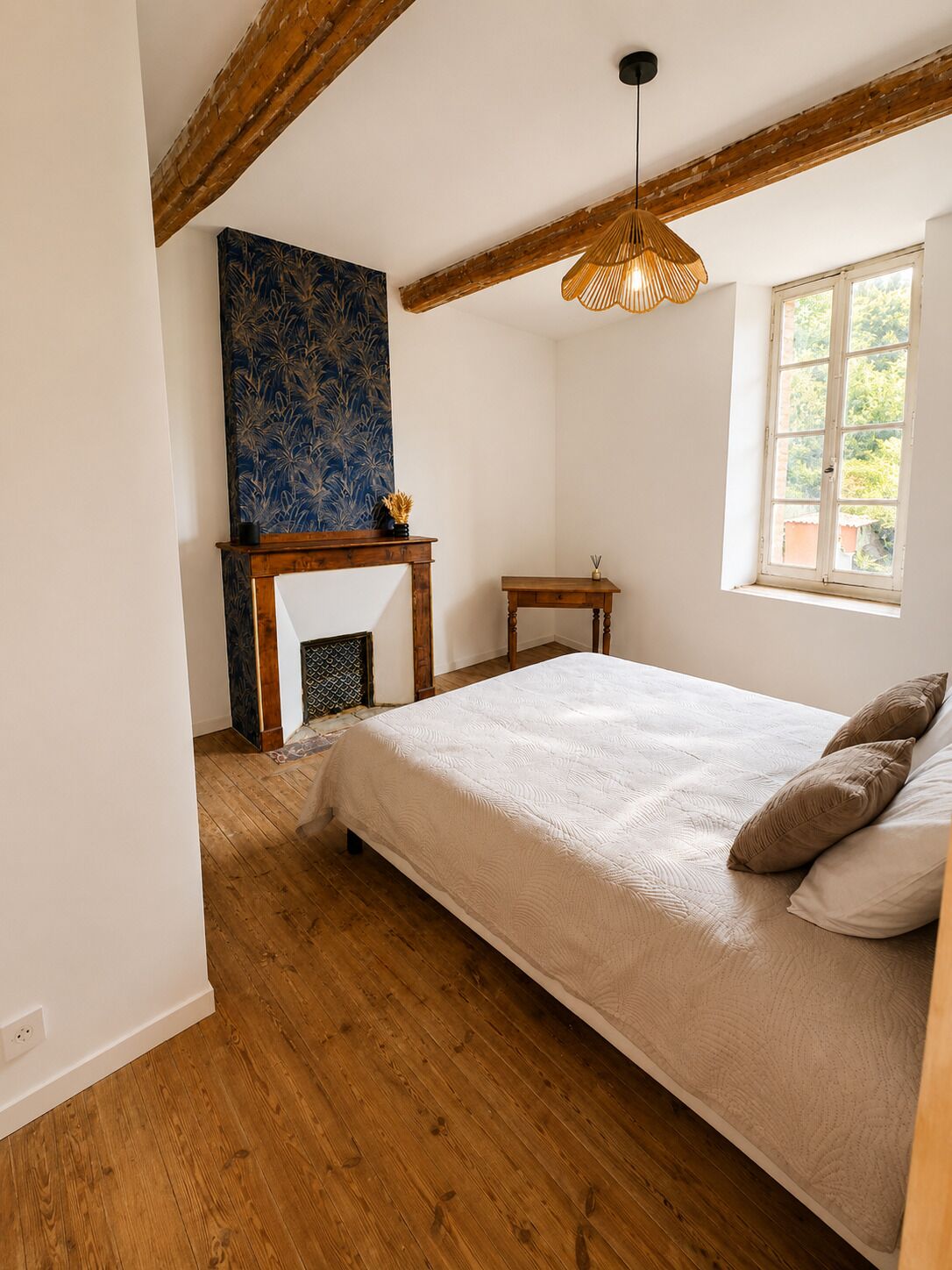 Un dormitorio con cama, piso de madera, chimenea, ventana y lámpara colgante.