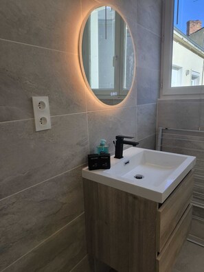Ein modernes Badezimmer mit einem weißen Waschbecken, einem schwarzen Wasserhahn und einem Spiegel mit Beleuchtung. Auf der Ablagefläche befinden sich zwei kleine Willkommensgeschenke.