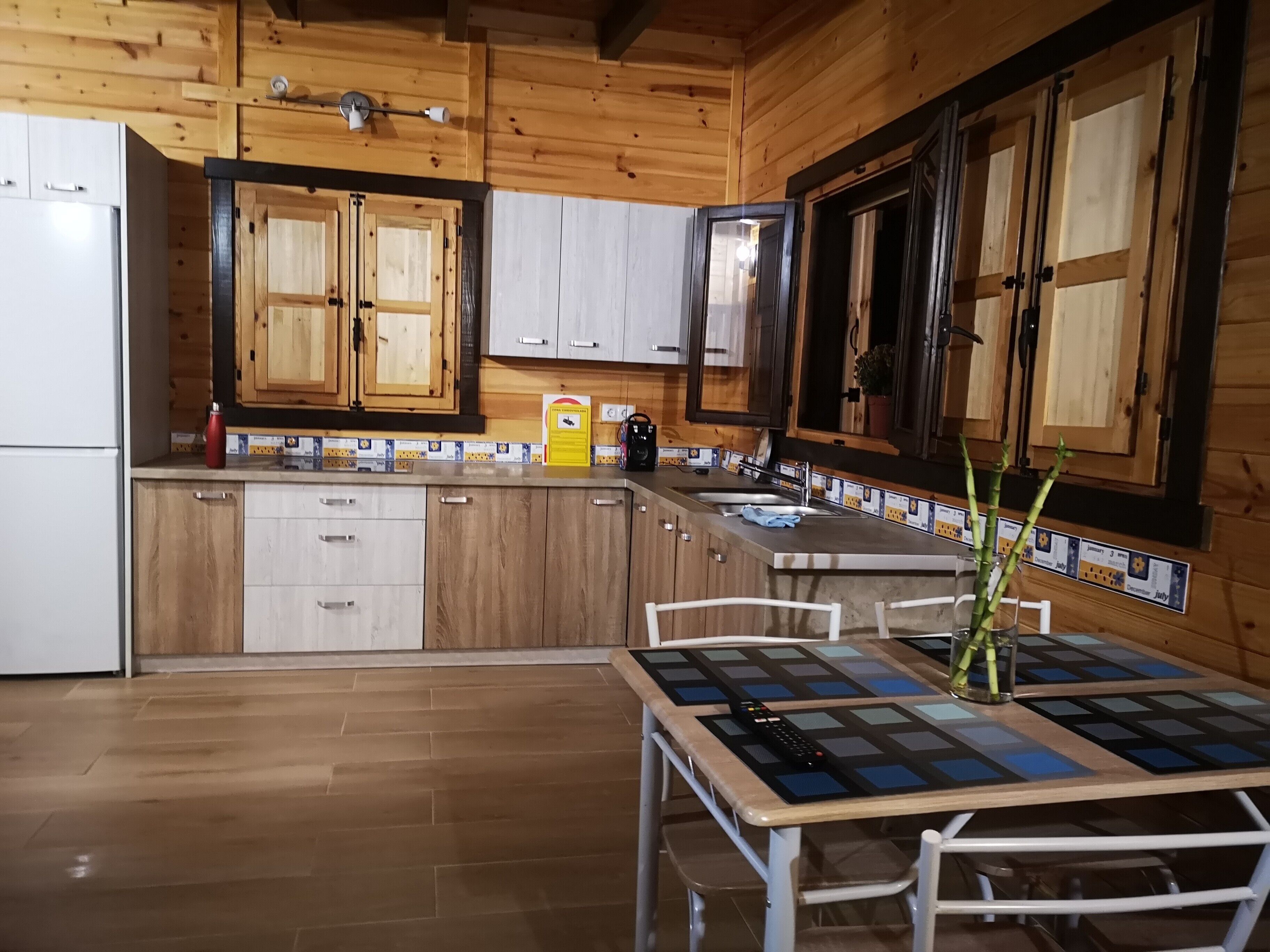 Une cuisine avec des meubles de rangement en bois, un réfrigérateur, un évier et une table recouverte d’une nappe à carreaux.