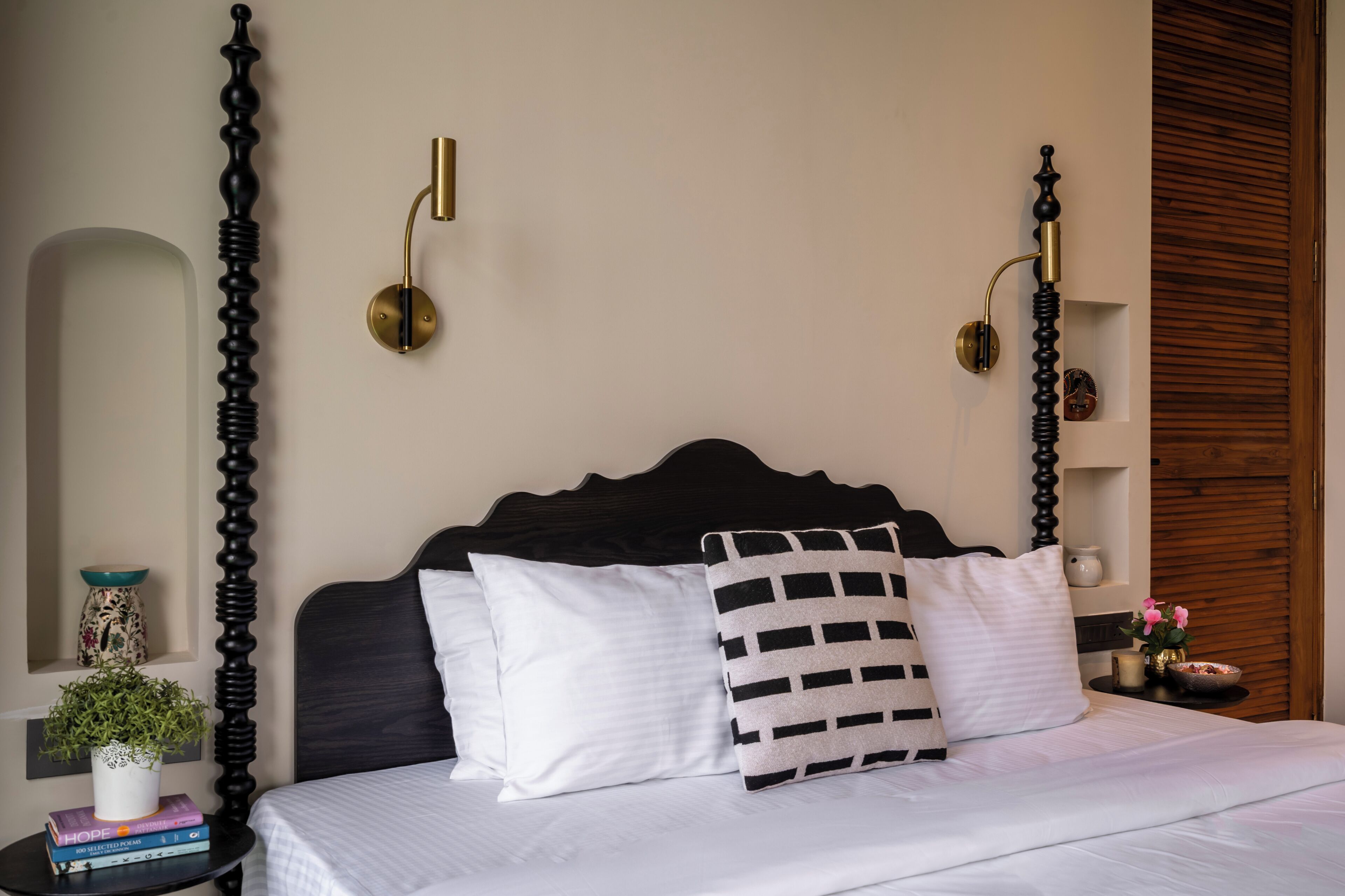 A neatly made bed with white and striped pillows, a bedside table with books and a plant, and a wall-mounted lamp.
