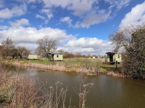 A tranquil riverside setting with a row of small, light-colored cabins, a wooden bench, and a grassy area. - Hop To It (Whitchurch)