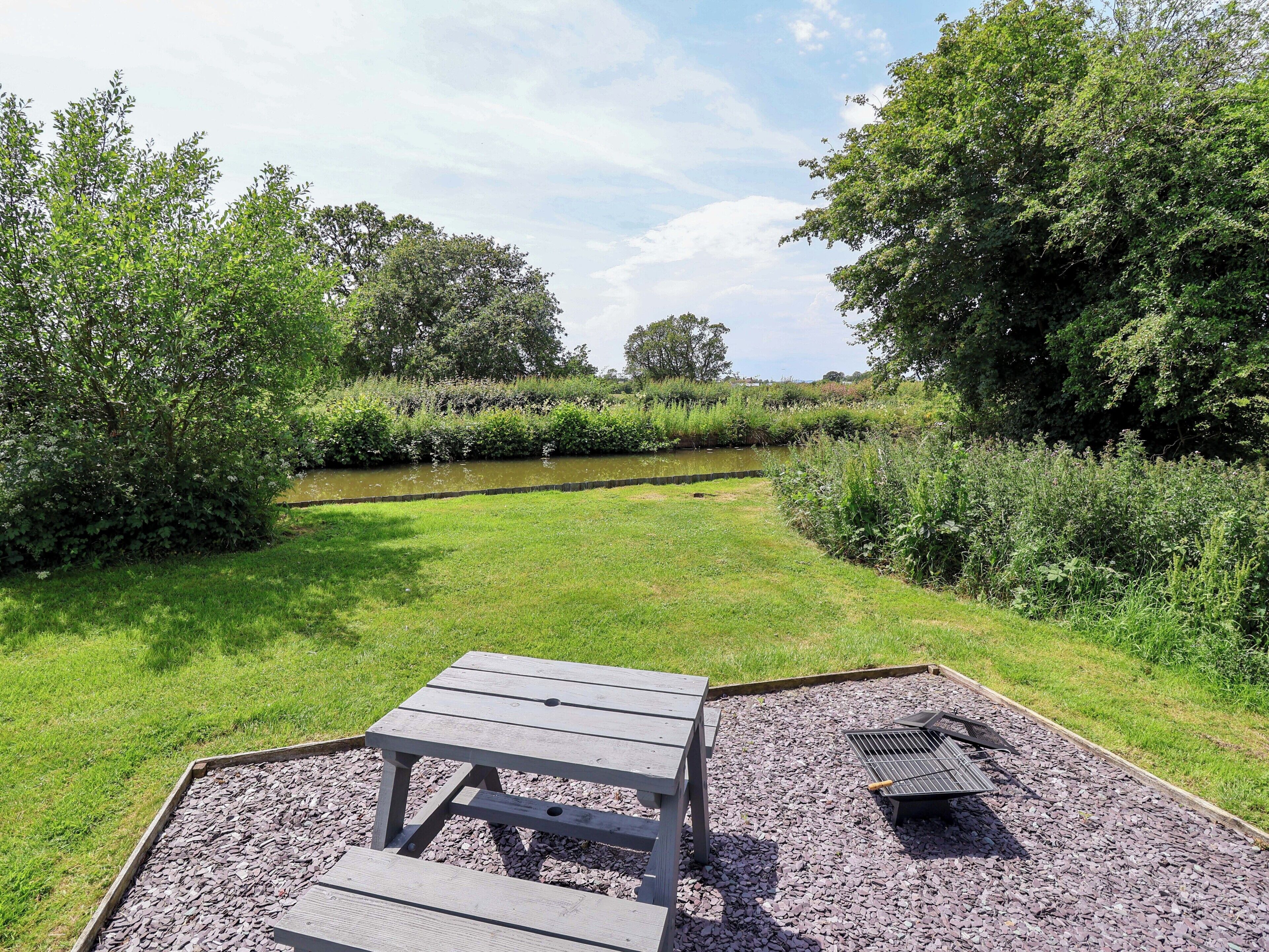 A picnic table with a grill, surrounded by gravel and grass, with a pond and trees in the background.