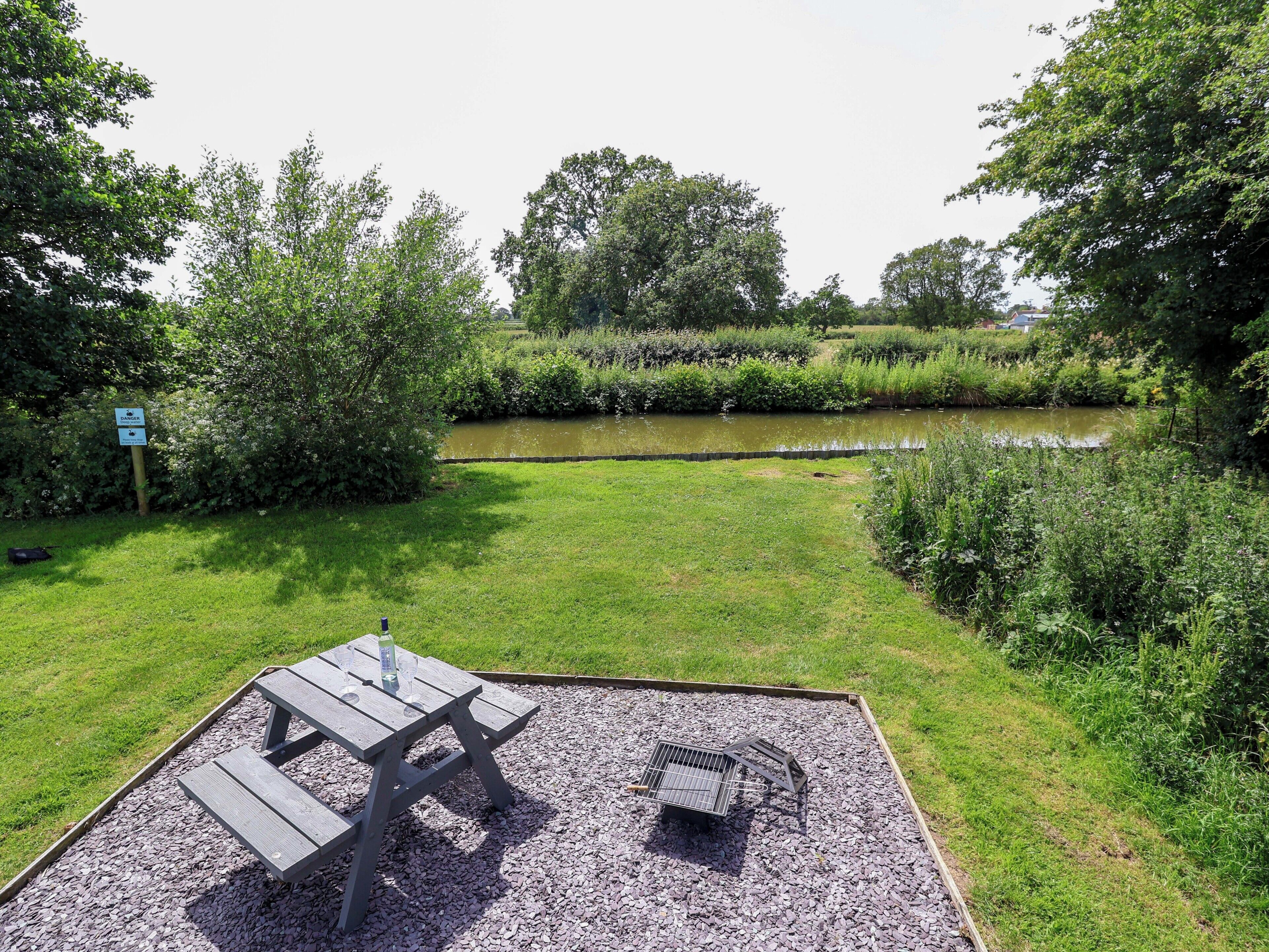 A picnic area with a table and benches, surrounded by gravel and grass, with a river and dense foliage in the background.