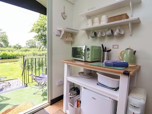 A kitchen with a green microwave, a kettle, and a coffee maker on the counter. There are shelves above holding cups and a welcome sign. - Hop To It (Whitchurch)