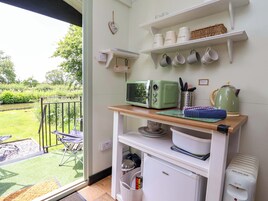 Una cocina con microondas verde, una tetera y una cafetera sobre la encimera. Hay estantes arriba con tazas y un cartel de bienvenida.