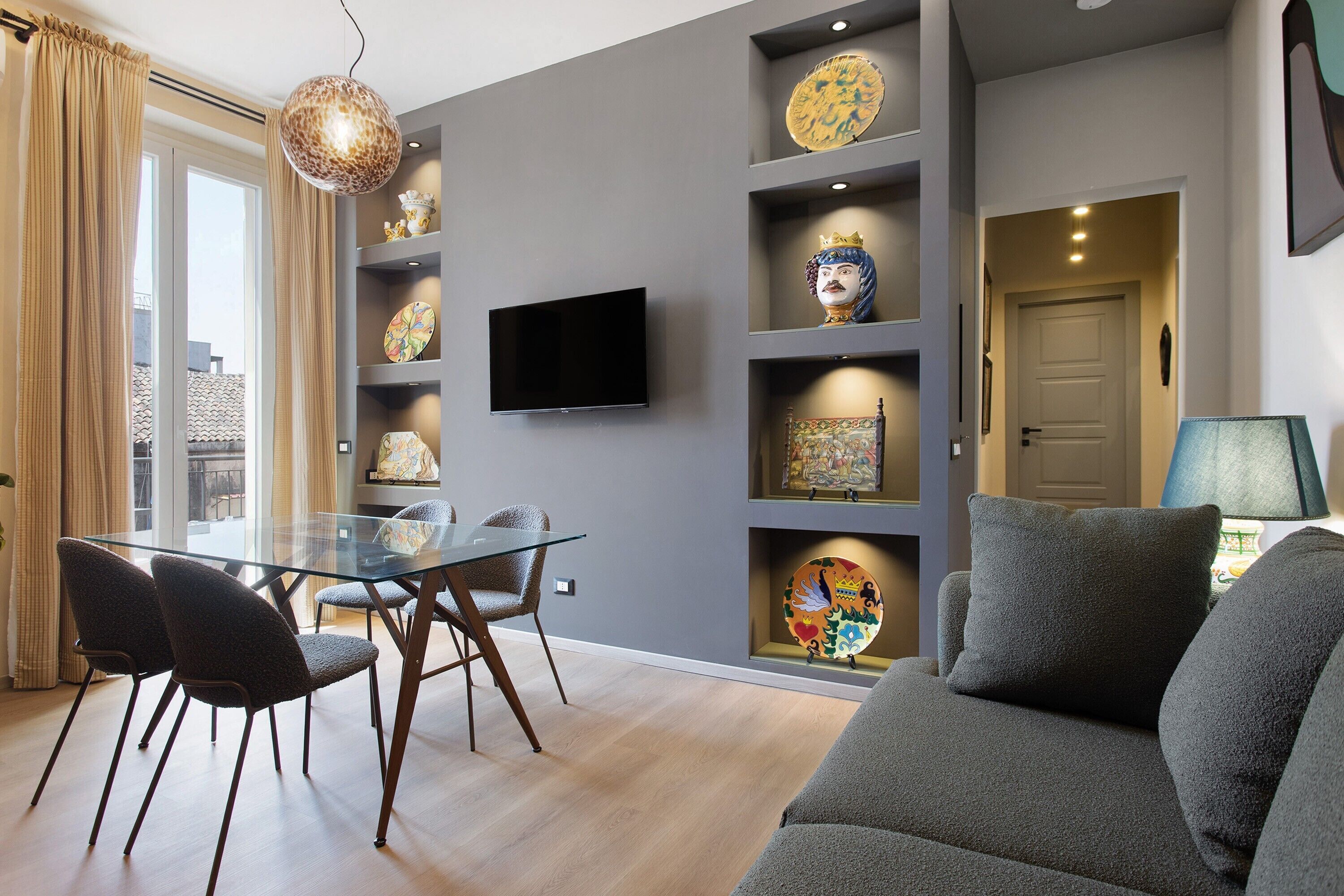 A modern living room with a glass dining table, grey sofas, and decorative items on shelves.