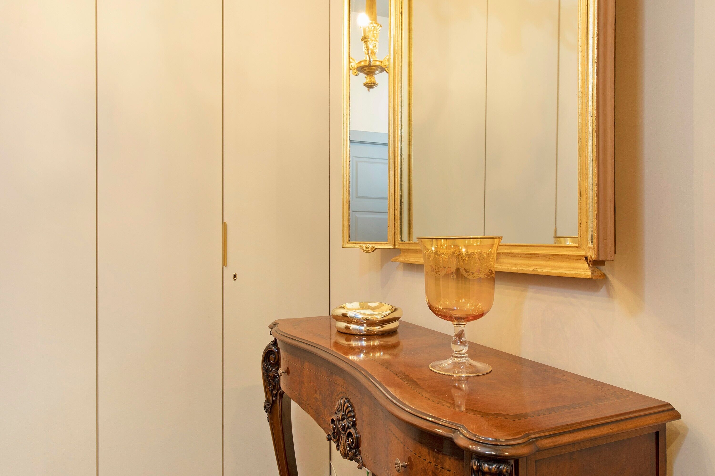 Una consola de madera con un florero de cristal y un plato decorativo, ubicada frente a un espejo con marco dorado.