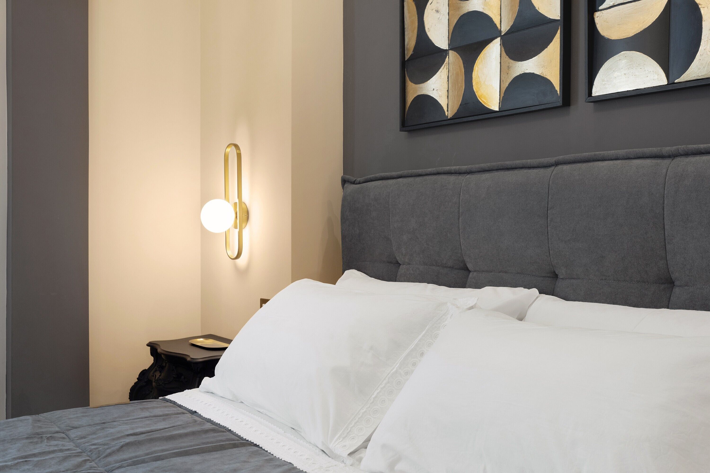 Een keurig opgemaakt bed met witte lakens, een grijs gestoffeerd hoofdbord, en een wandlamp.