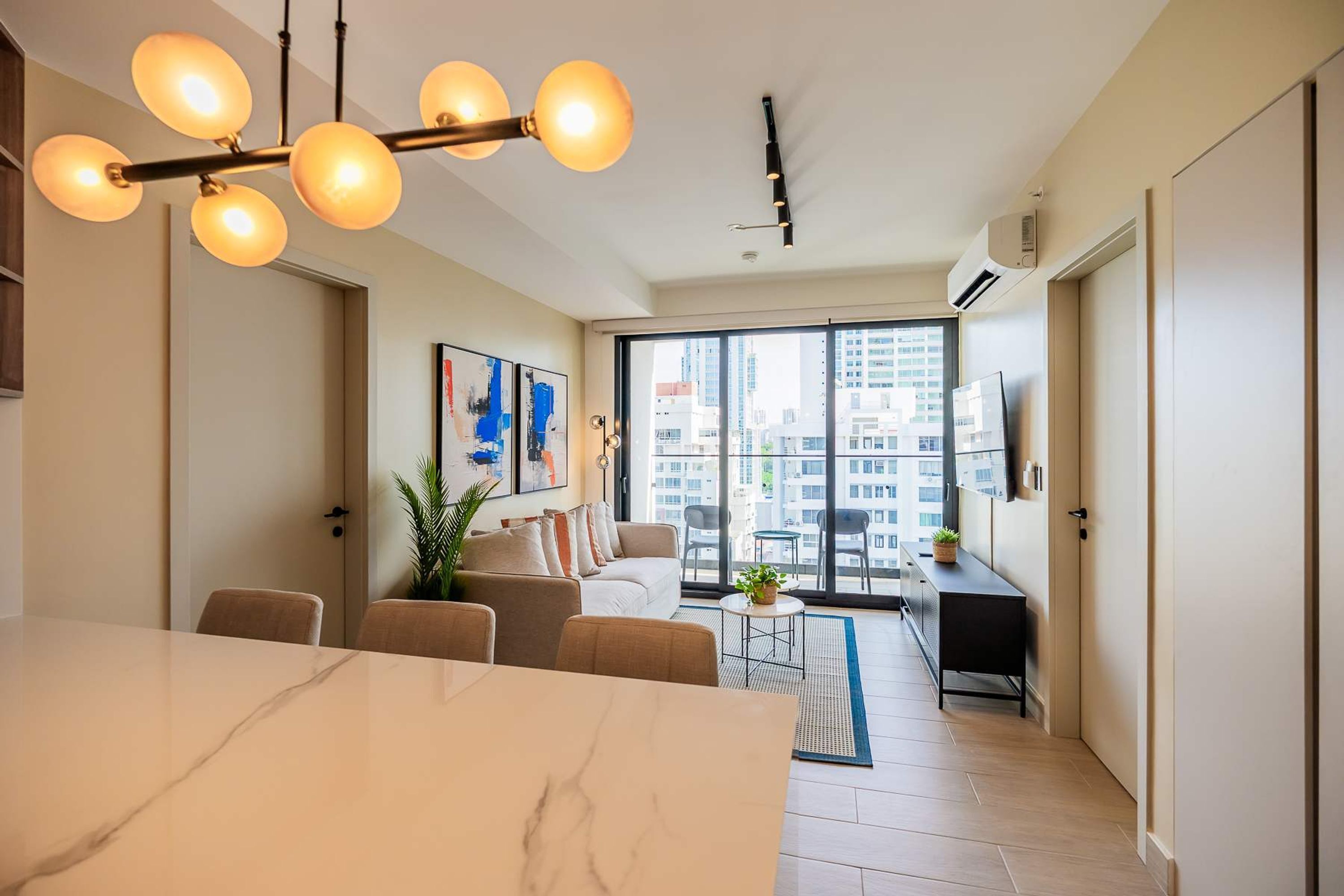 A modern living room with a large window, a dining area with a marble table, and a ceiling light fixture.
