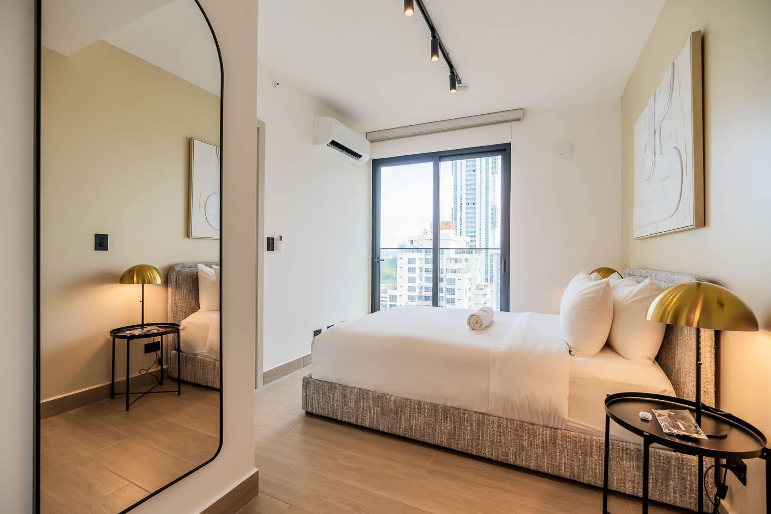 A modern hotel room with a large bed, a small side table, a mirror, and a window with a city view.