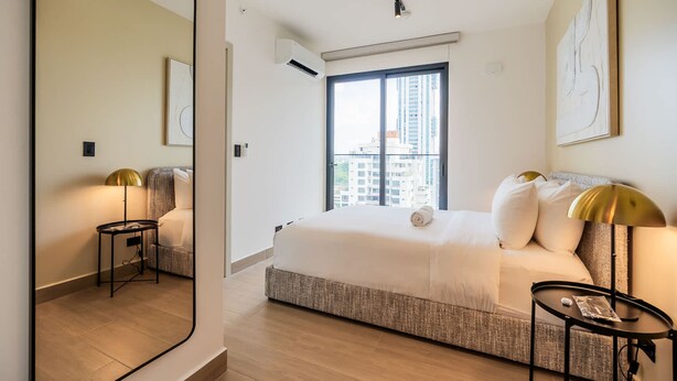 A modern hotel room with a large bed, a small side table, a mirror, and a window with a city view. - w Amazing 2BR w Balcony AC in San Francisco (Panama City)