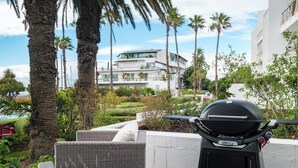 En terrasse med grill, fletmøbler og udsigt til en moderne bygning med swimmingpool.