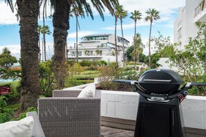 A patio with a grill, wicker furniture, and a view of a modern building with a pool. - Safe 2 Bed With View in The Waterclub Bella Marina (Cape Town)