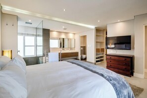 A modern hotel bedroom with a large bed, a vanity with a mirror, a television, and a walk-in closet. - Safe 2 Bed With View in The Waterclub Bella Marina (Cape Town)