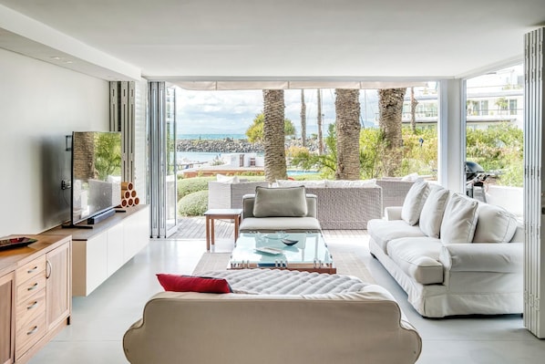 A modern living room with a sofa, coffee table, and TV, overlooking a pool and palm trees. - Safe 2 Bed With View in The Waterclub Bella Marina (Cape Town)
