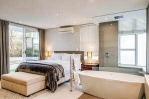 A modern hotel room with a large bed, a separate sofa, a bathtub, and a sliding glass door leading to a balcony. - Safe 2 Bed With View in The Waterclub Bella Marina (Cape Town)
