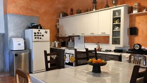 A kitchen with white cabinets, a stove, a refrigerator, and a table with a fruit bowl. - Vathia Tower - Stone Mani Heaven Near the Beach (East Mani)