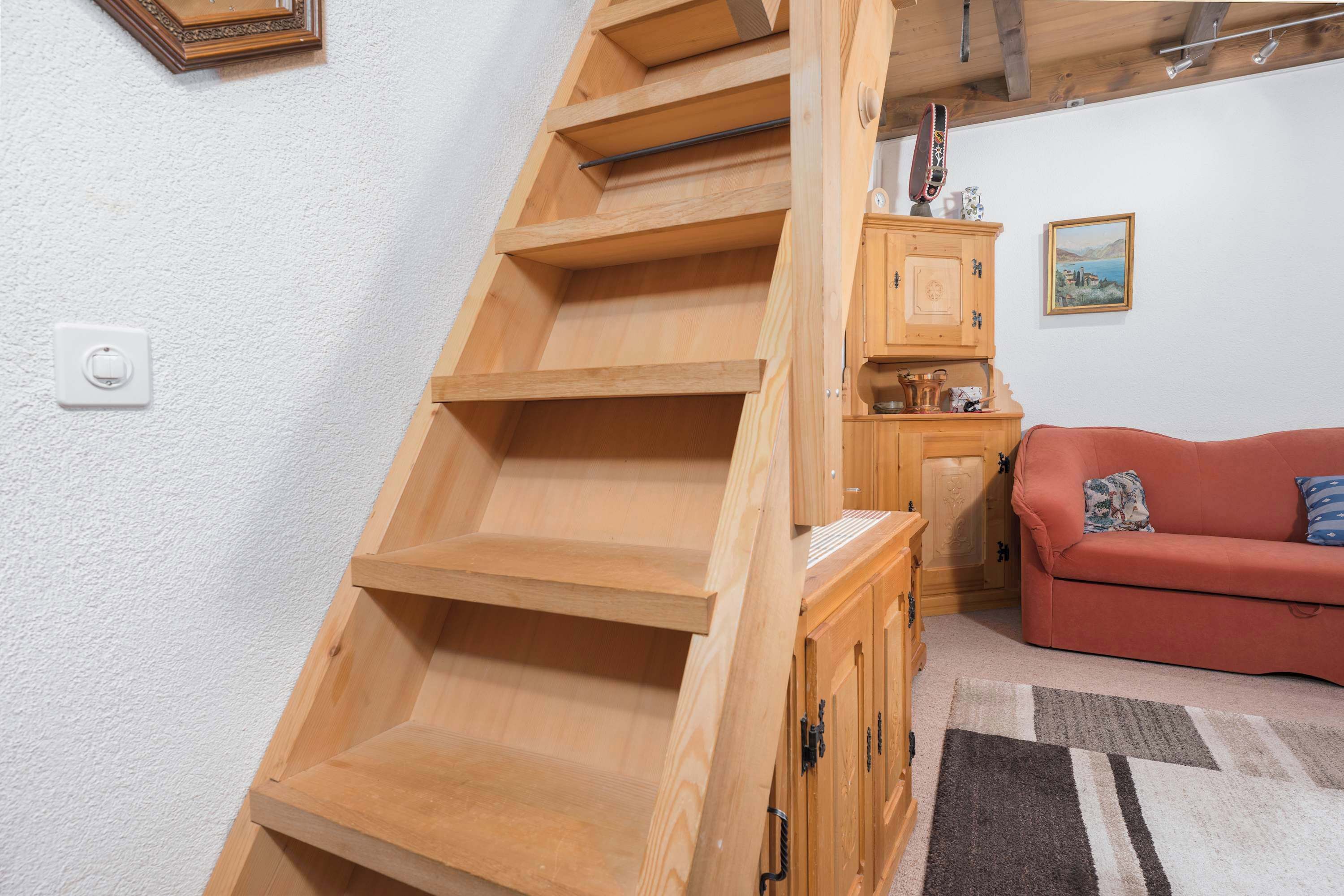 Un escalier en bois avec une rampe, un canapé rouge et un meuble surmonté d’une horloge.