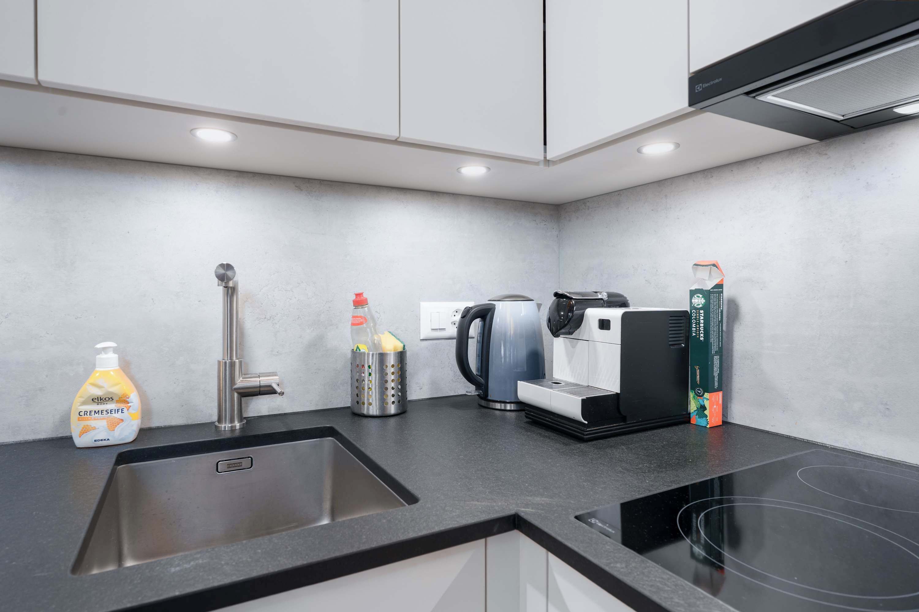 A modern kitchen with a sink, a kettle, a coffee machine, and a bottle of Elkos body cream.