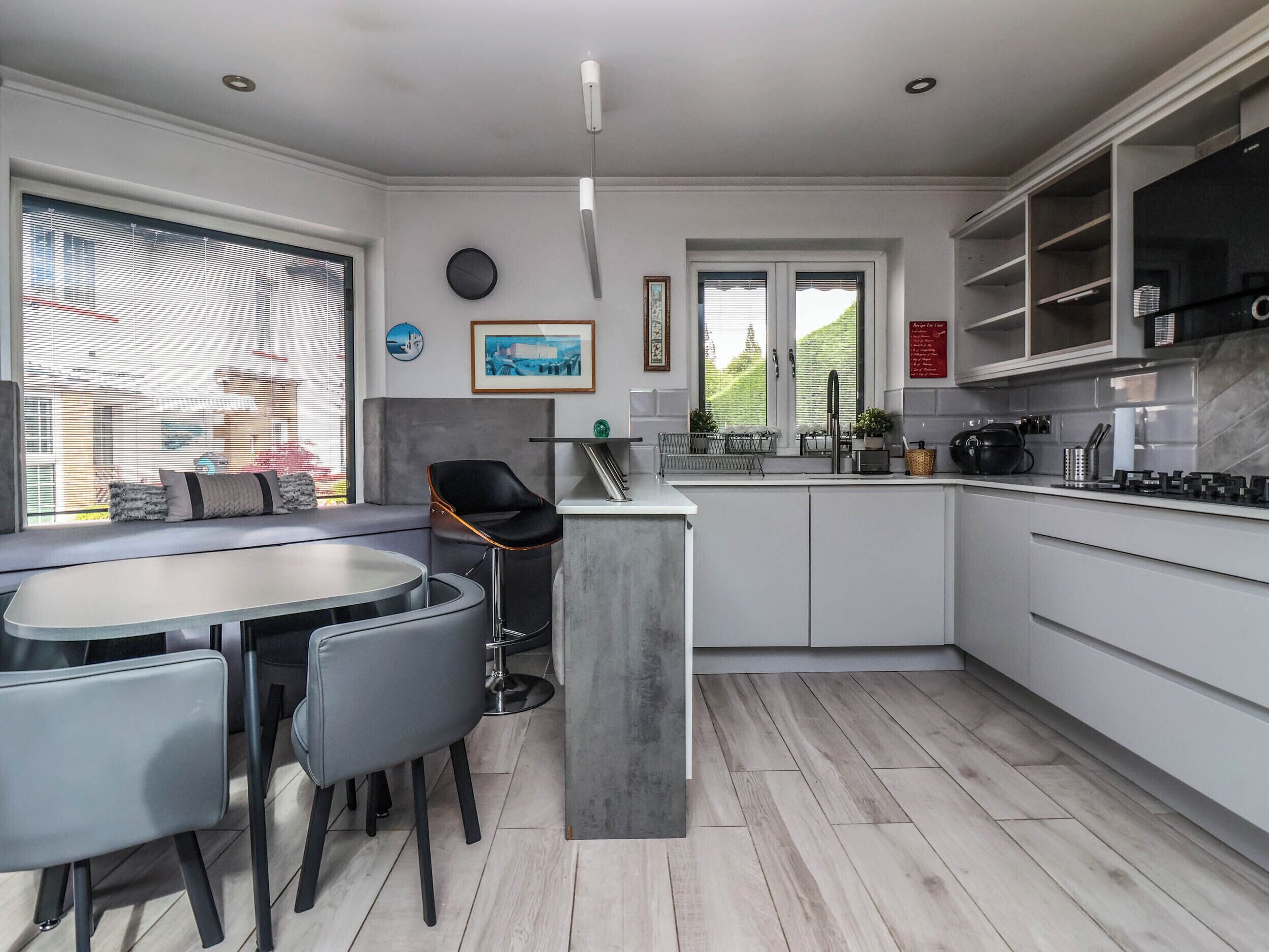 A modern kitchen with a dining area, featuring a central island, grey cabinetry, and a view of the outdoors.