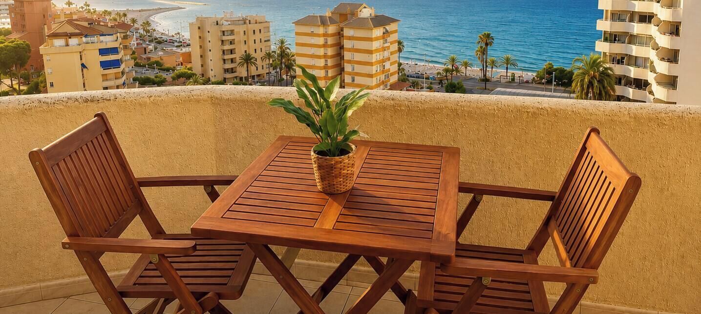 Ein Holztisch mit Stühlen auf einem Balkon, Blick auf Strand und Gebäude.