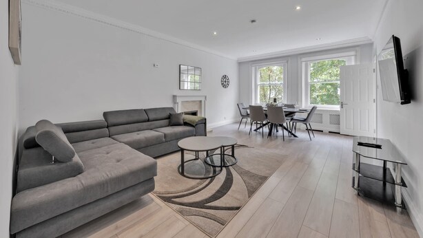 A modern living room with a grey sectional sofa, a round coffee table, and a dining area with a glass table and chairs. - Luxury 2-bedroom Apartment Next to Harrods (London)