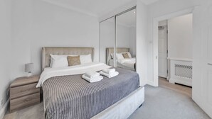 A modern bedroom with a large bed, a nightstand, a mirror, and a door leading to another room. - Luxury 2-bedroom Apartment Next to Harrods (London)