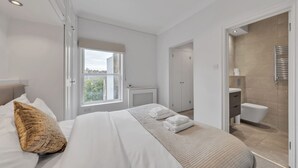 A modern hotel room with a large bed, a window, a bathroom, and a view of the city. - Luxury 2-bedroom Apartment Next to Harrods (London)