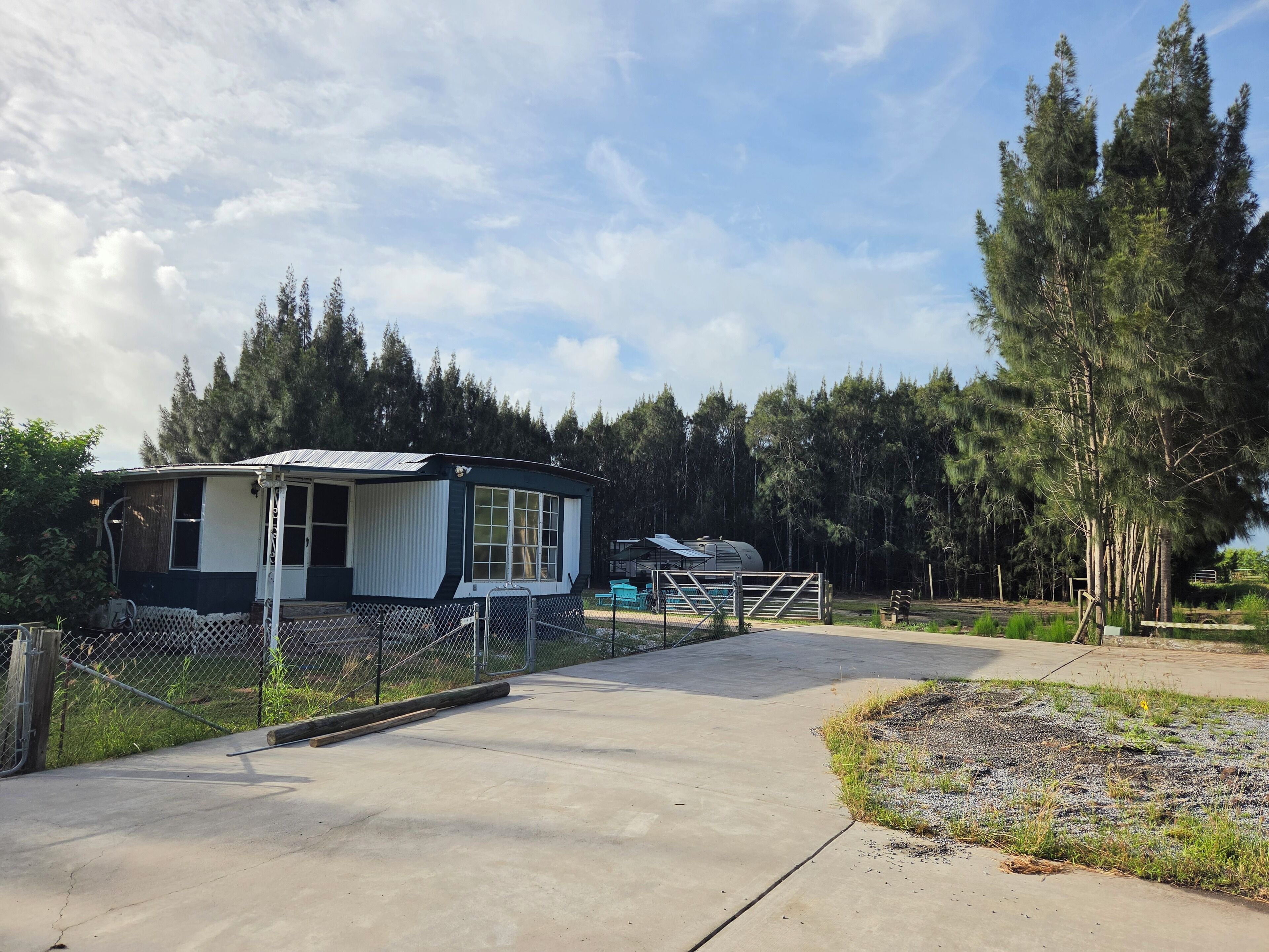 A mobile home with a fenced yard, a concrete driveway, and a forested backdrop.