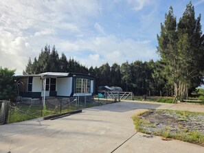 Una casa móvil con patio cercado, camino de hormigón y un paisaje boscoso de fondo.
