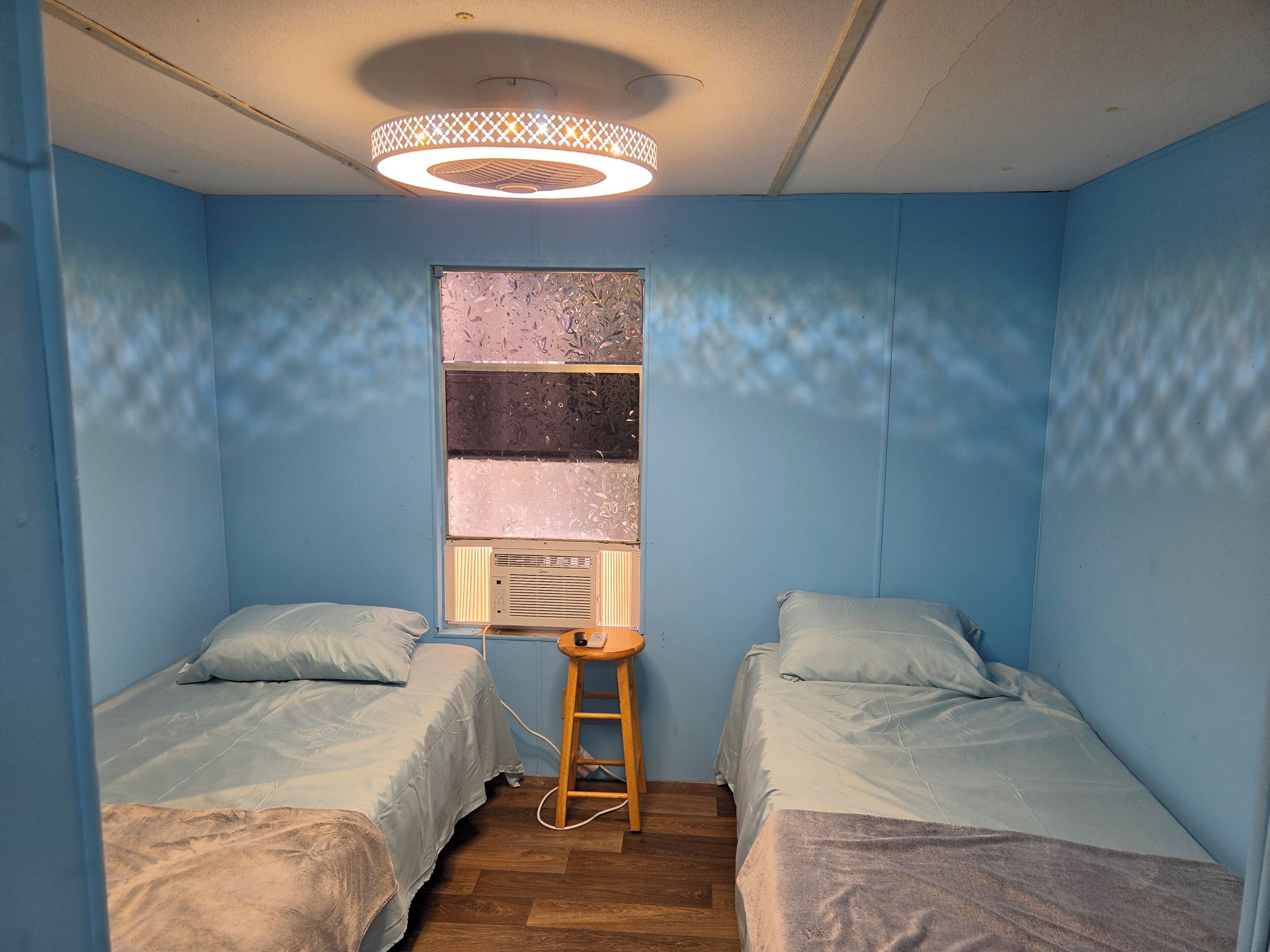 A small room with two beds, a window with a decorative pattern, a wooden stool, and a ceiling light fixture.