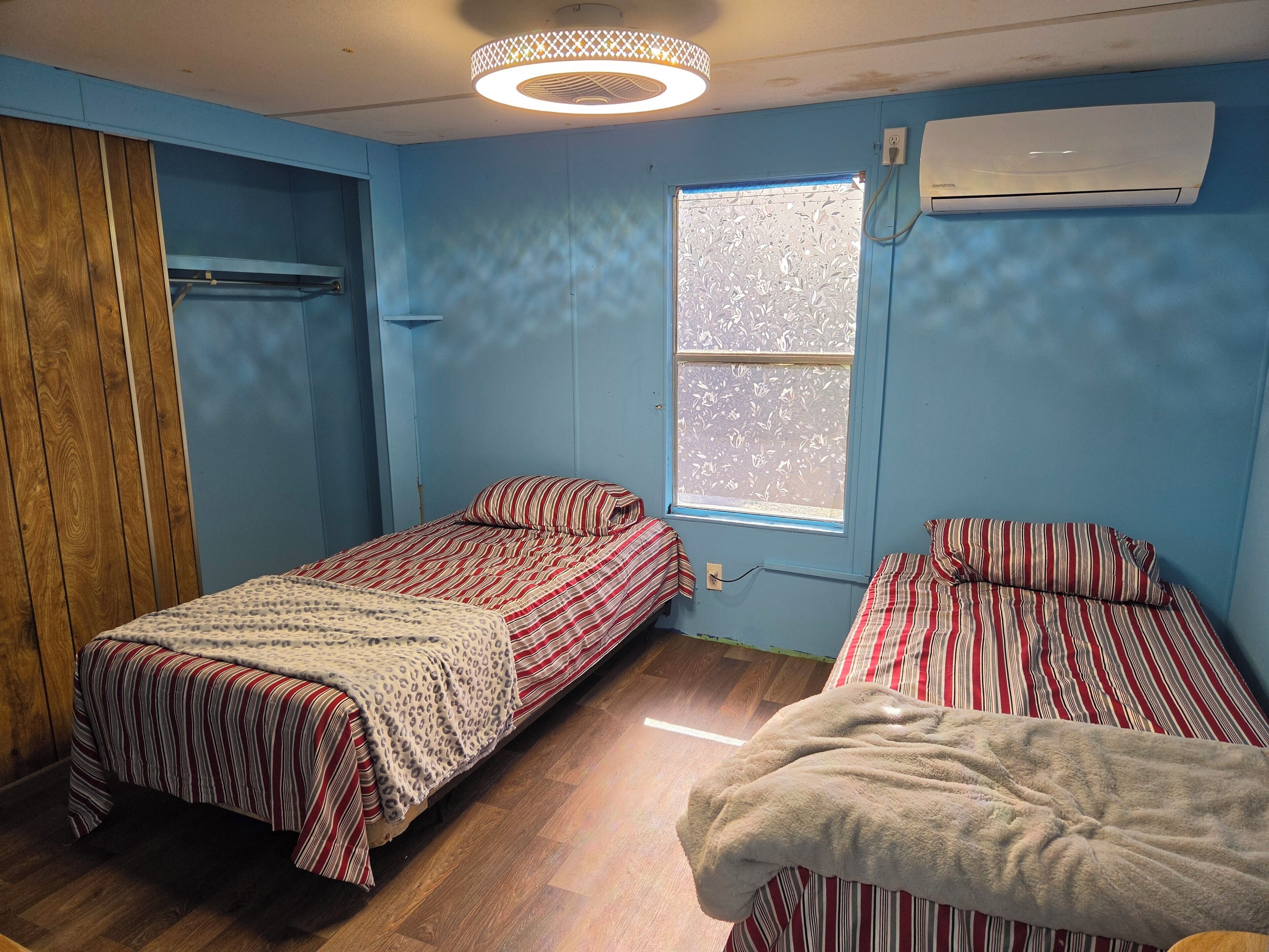 Een kamer met twee bedden, een raam met bloemprint rolgordijnen, een airconditioning en een houten kledingkast.